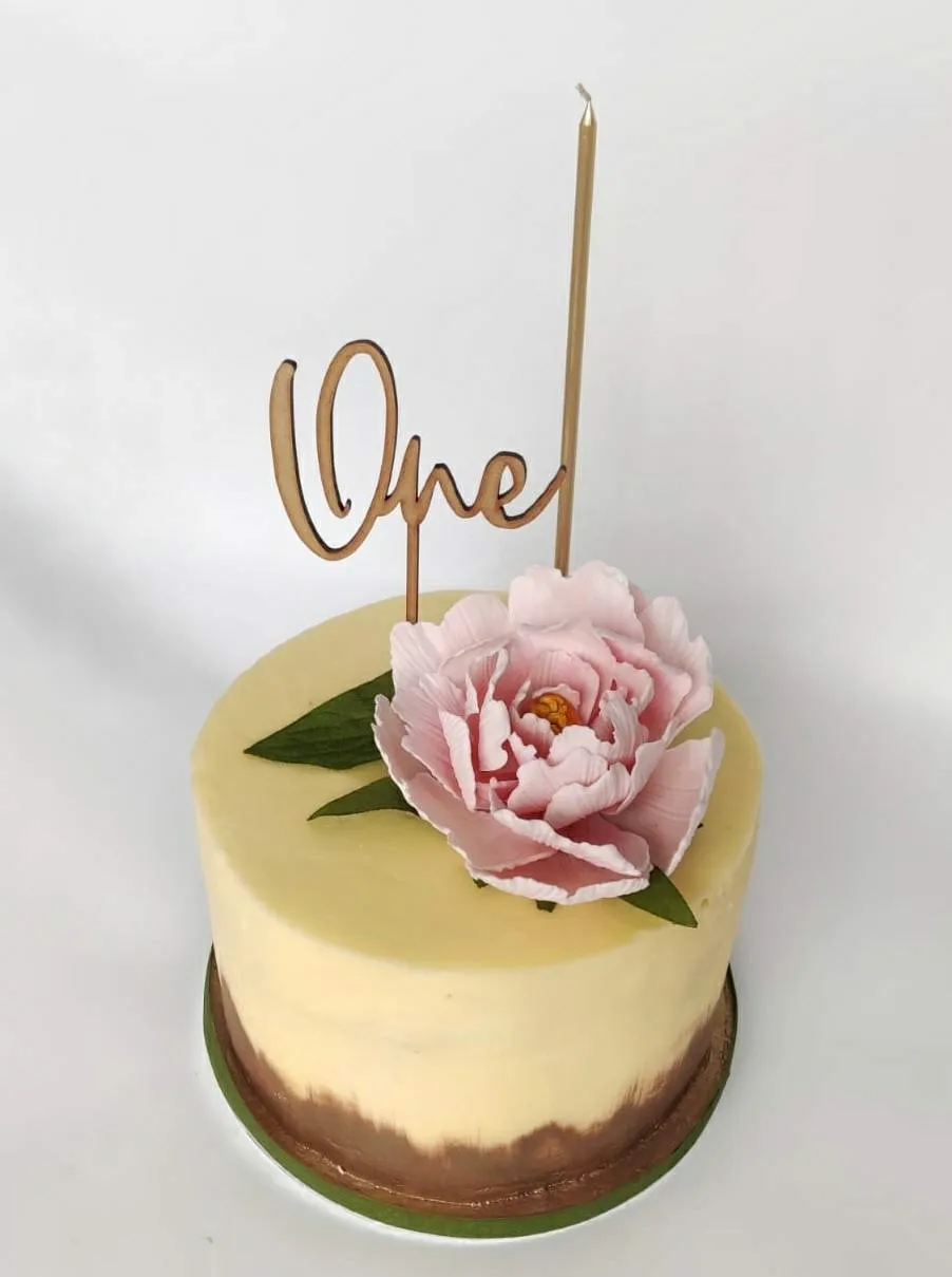 Elegant first birthday cake with smooth cream buttercream, rose gold base, and a large pink sugar peony