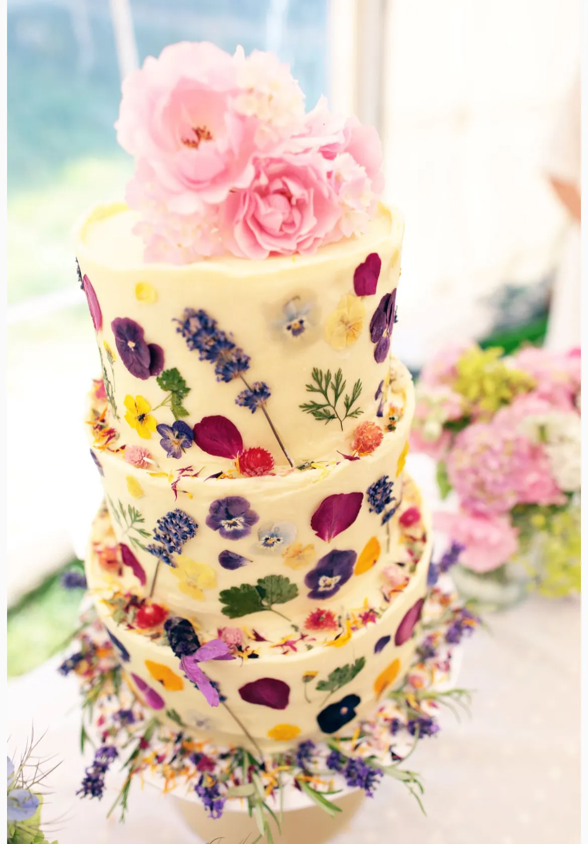Four-tier cake with vibrant pressed edible flowers in purple, yellow, burgundy, and pink creating a painted effect