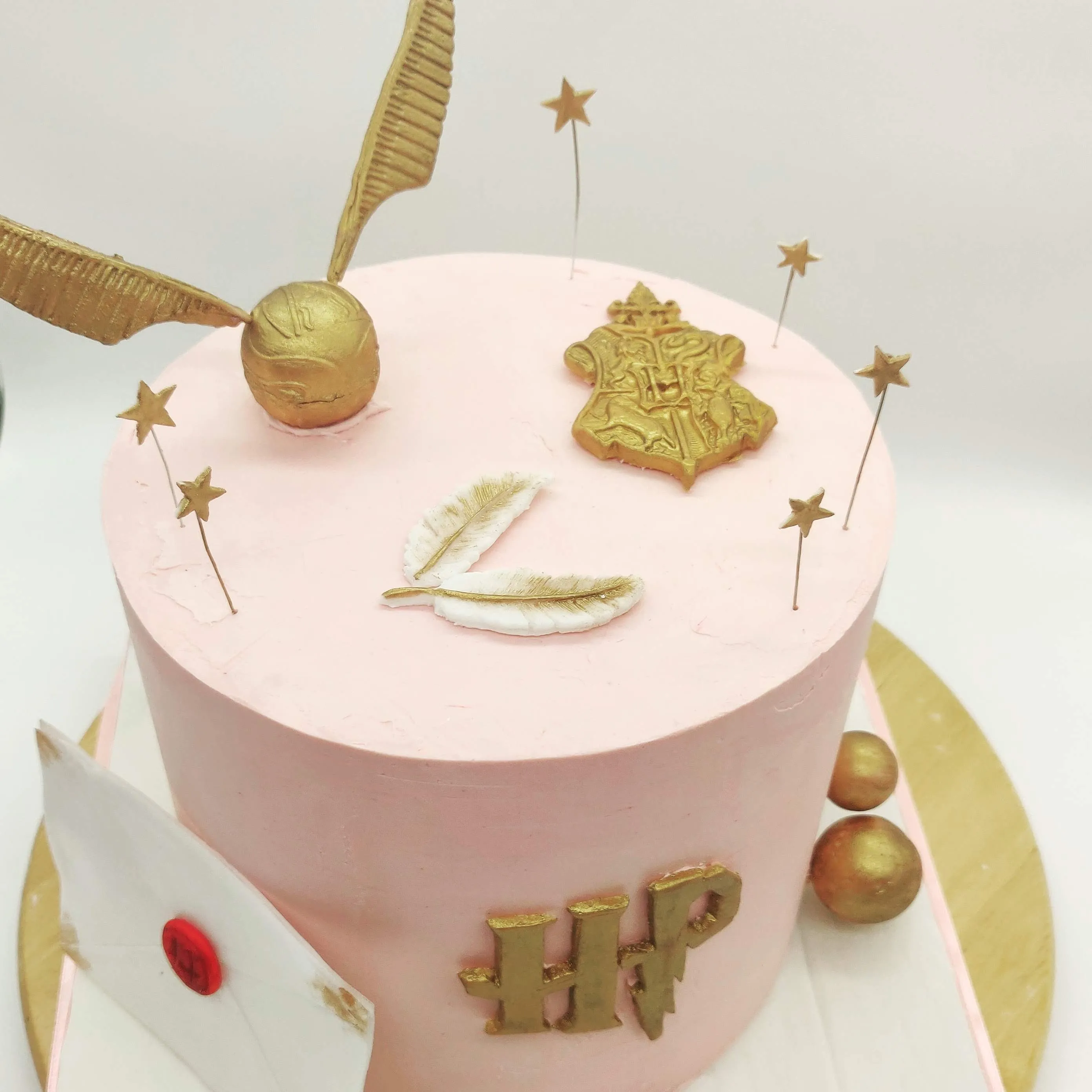 Pink Harry Potter birthday cake with gold fondant Golden Snitch, Hogwarts crest, white quill and gold stars
