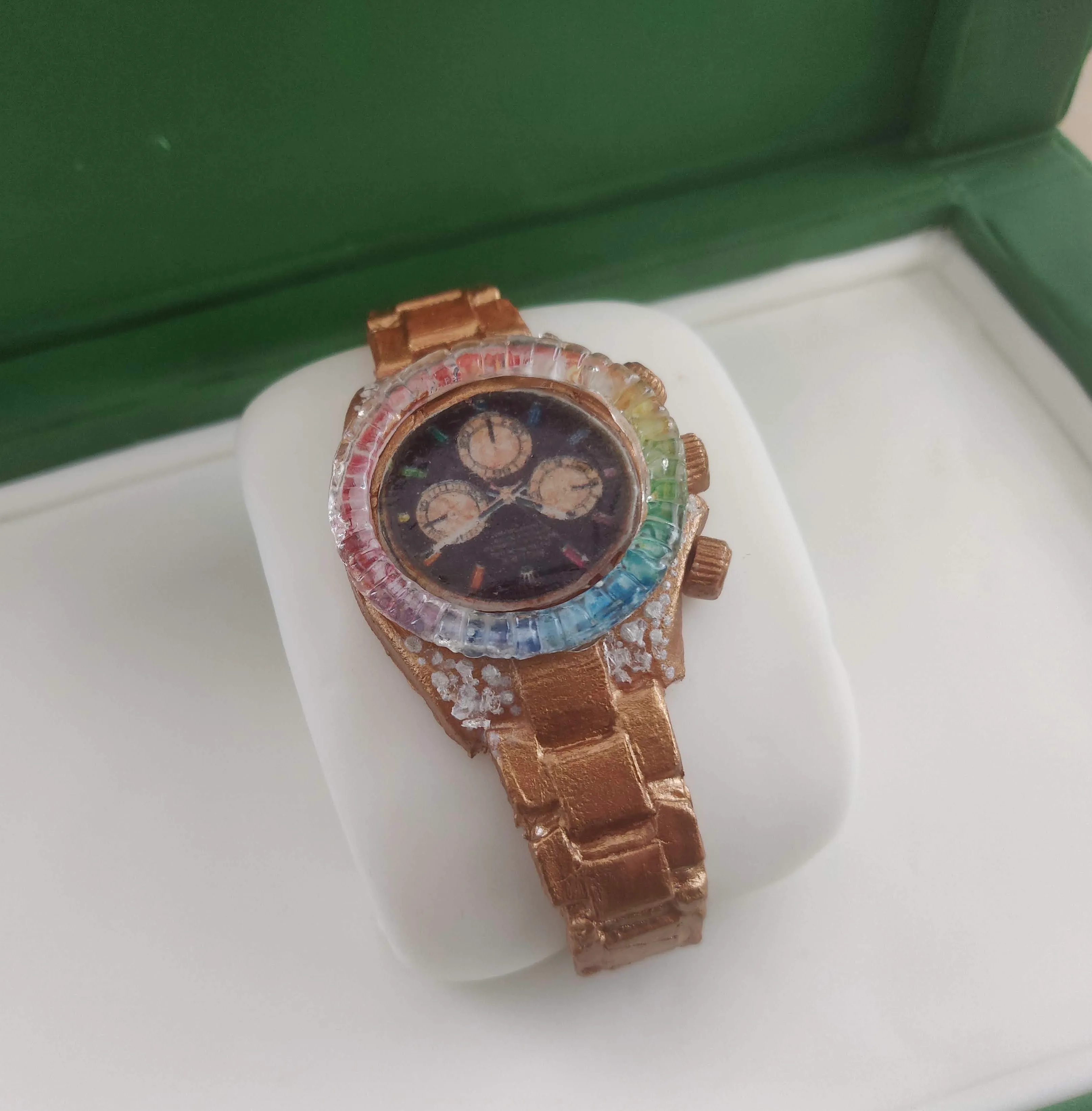 Sculpted gold luxury watch with rainbow-coloured bezel resting on a white cushion inside a green presentation box