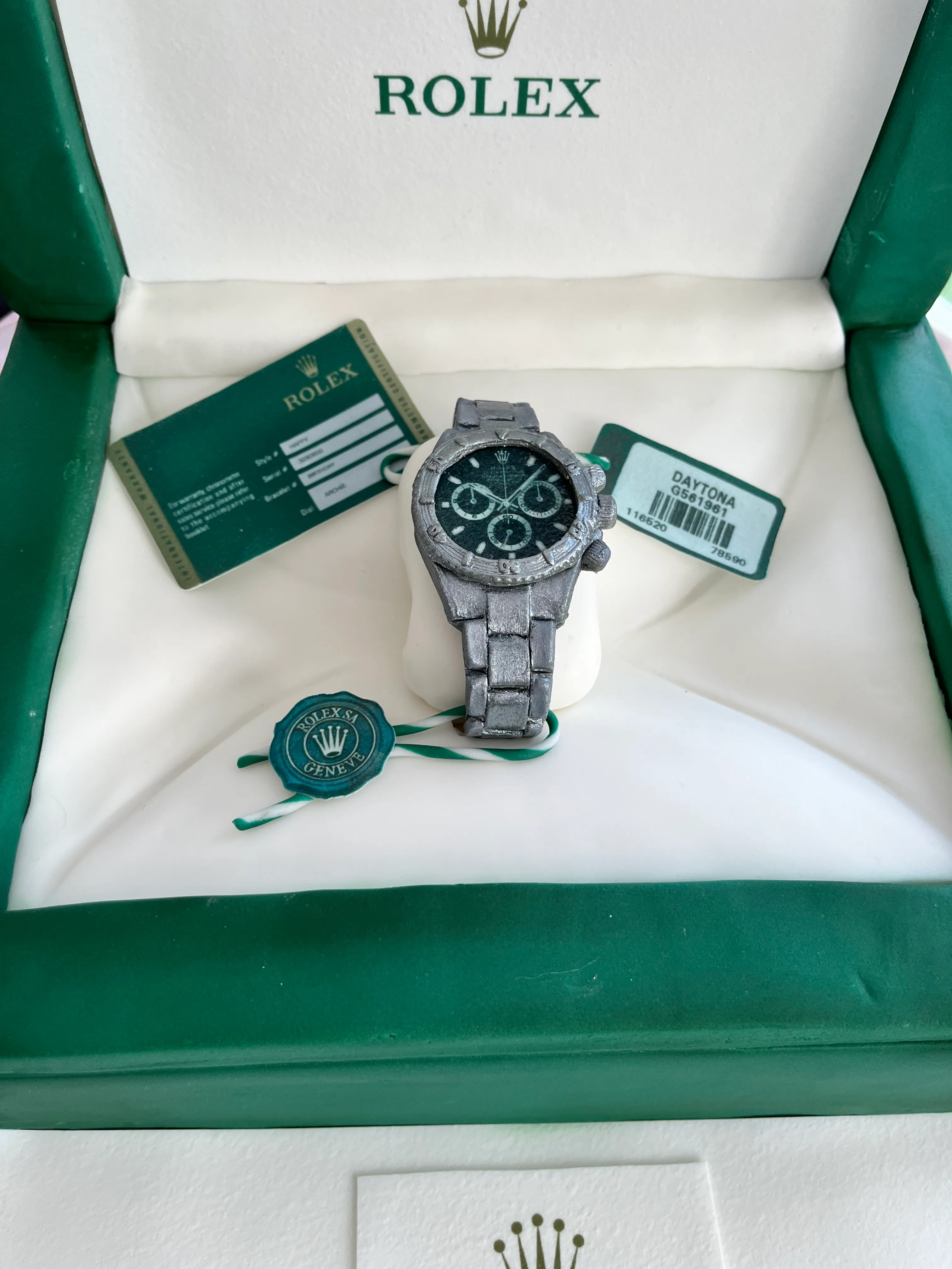 Close-up of sculpted silver Rolex Daytona watch with warranty card and wax seal on white cushion