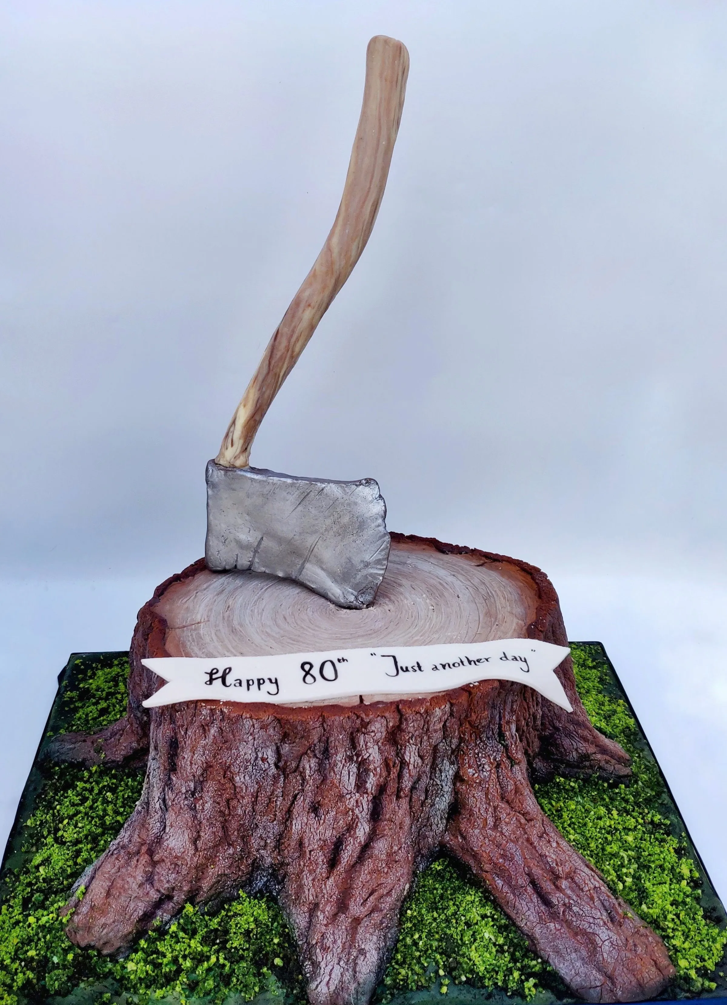 Realistic tree stump cake with detailed bark texture, silver axe embedded in top, and moss at the base