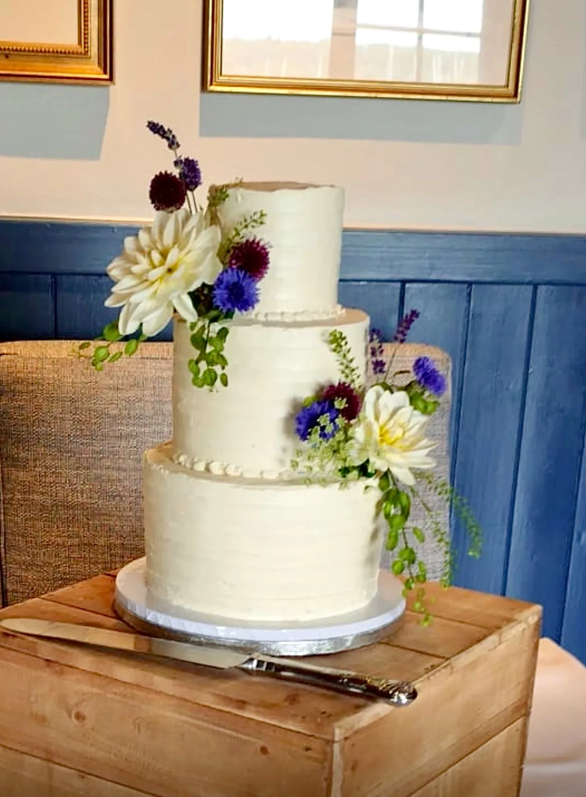 Three-tier white buttercream cake with white dahlias, purple cornflowers, and trailing greenery