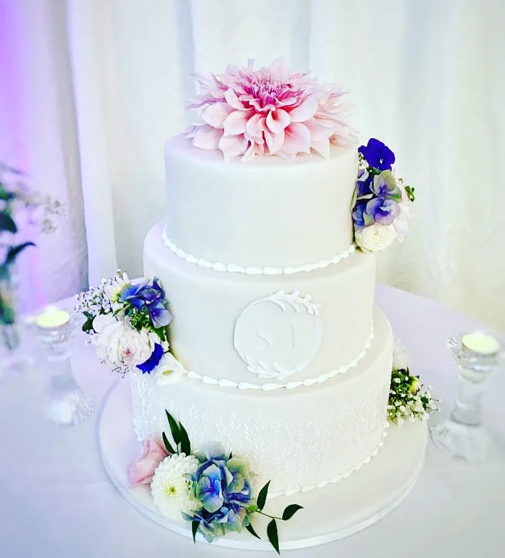 Three-tier white cake decorated with a pink peony, blue delphiniums, baby's breath, and gold beaded details