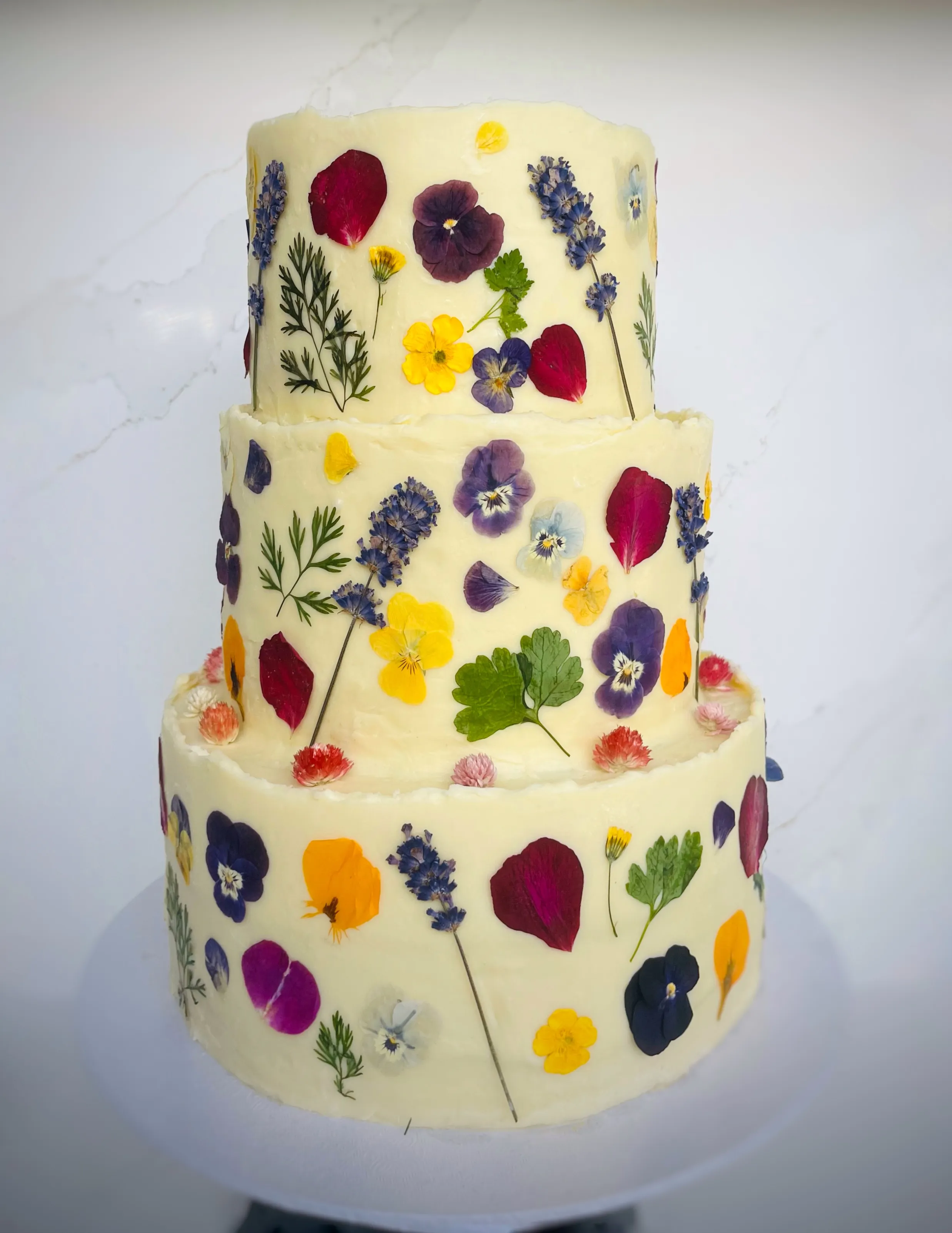 Three-tier cream cake decorated with colourful pressed edible flowers including purple, yellow, and pink pansies